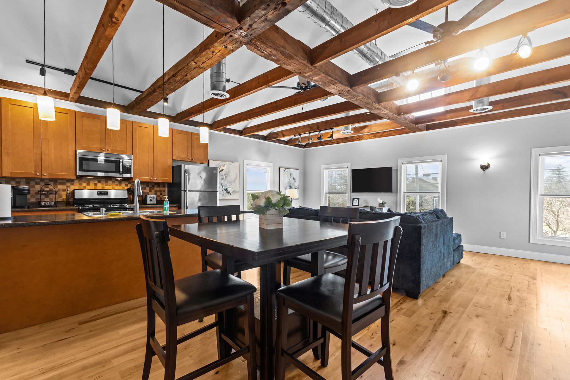 Open-concept living space with kitchen, dining area, and living room. Exposed wooden beams, wood floors, and neutral walls.