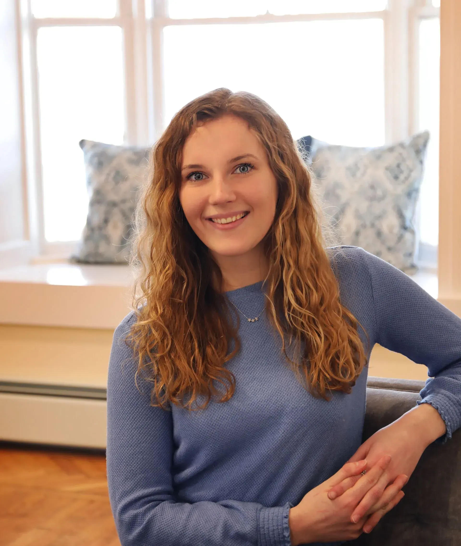 Woman with curly brown hair smiles, sitting on a couch. Blue sweater, window with pillows in background.