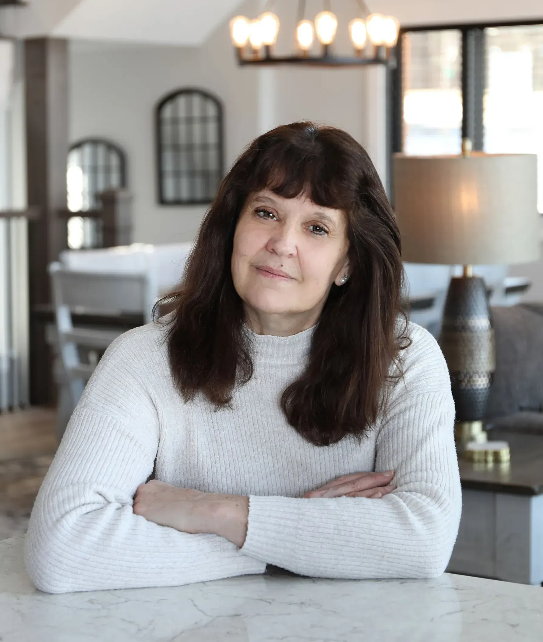 Woman with dark hair and bangs, arms crossed, smiling slightly, in a modern, well-lit home.