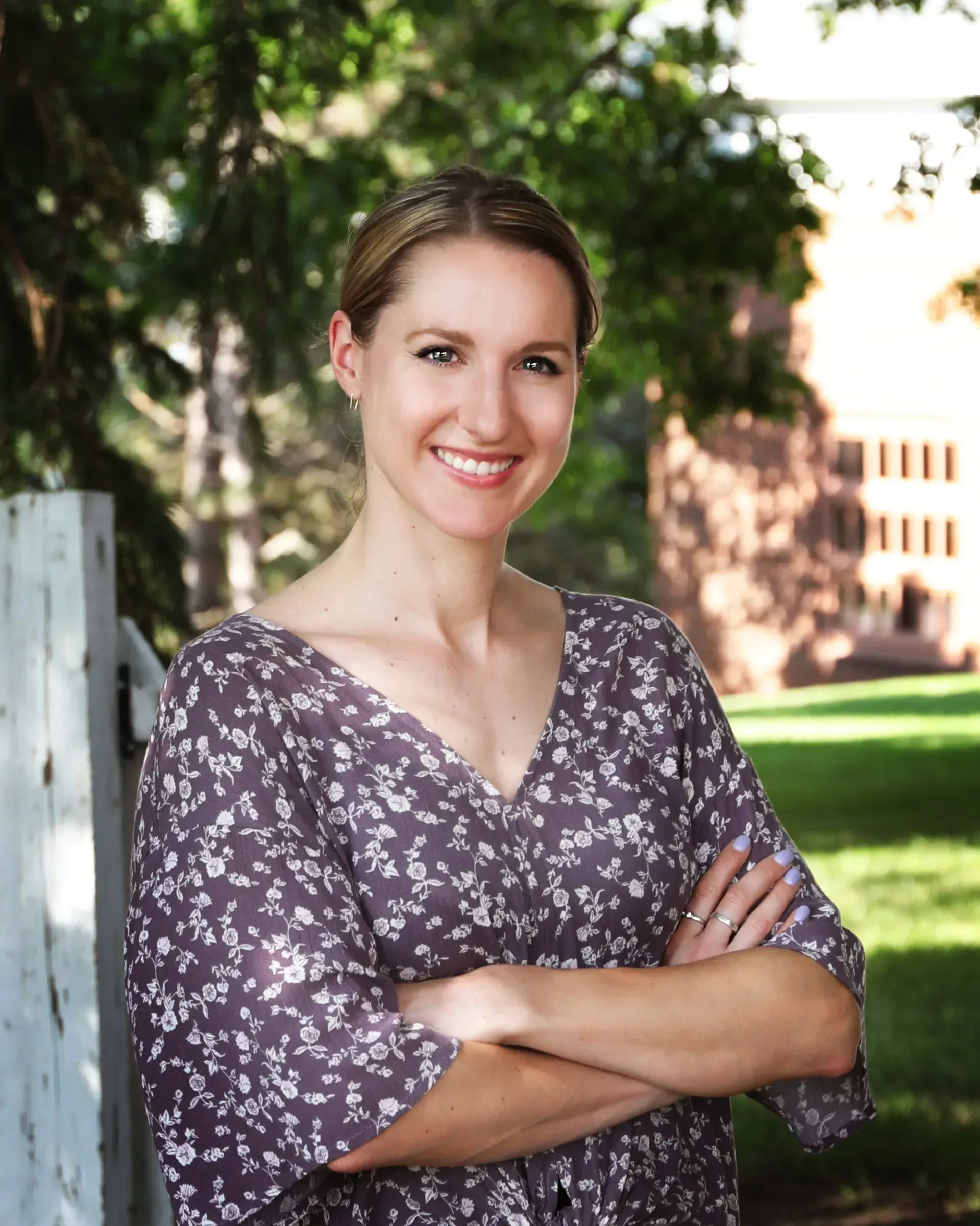 Woman smiling with arms crossed, standing near a white fence and greenery; brick building in the background.