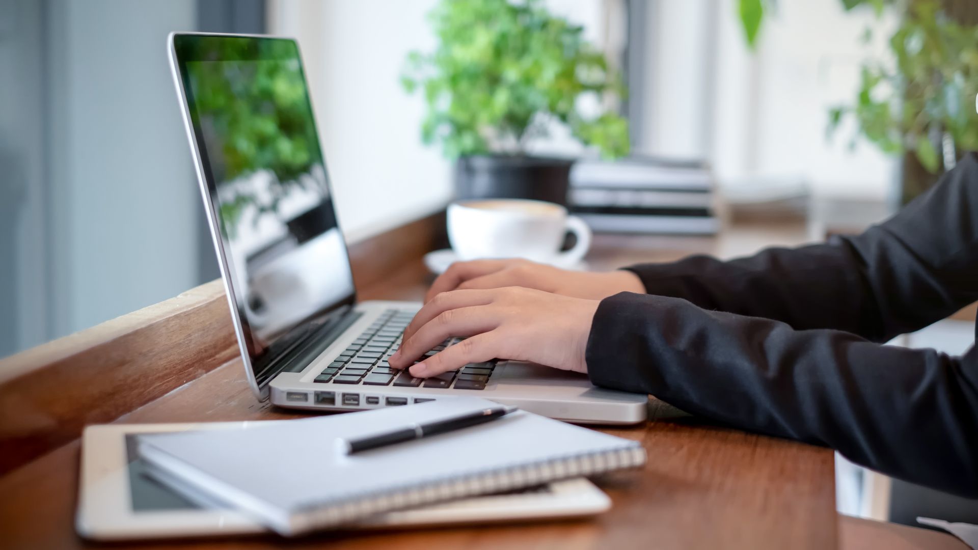 Person typing on a laptop at a wooden table, with a notebook, pen, and coffee cup nearby.
