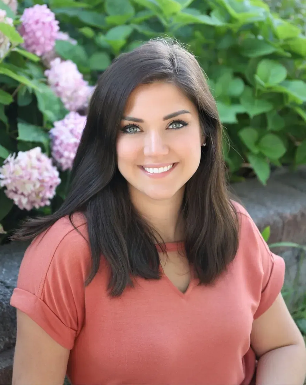 Woman with dark hair smiles, wearing coral top, posing outdoors with pink flowers in background.