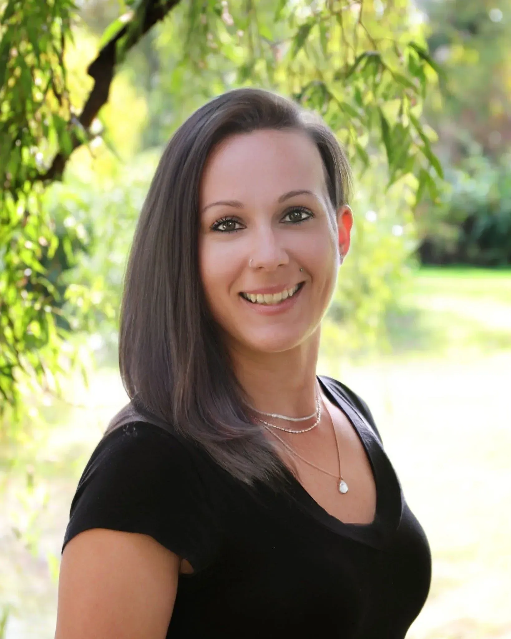 Woman with dark hair smiles outdoors, wearing a black top and jewelry, blurred green background.
