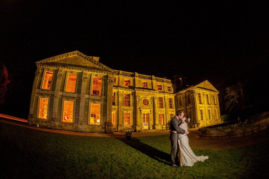 Couple kissing in front of a brightly lit mansion at night.