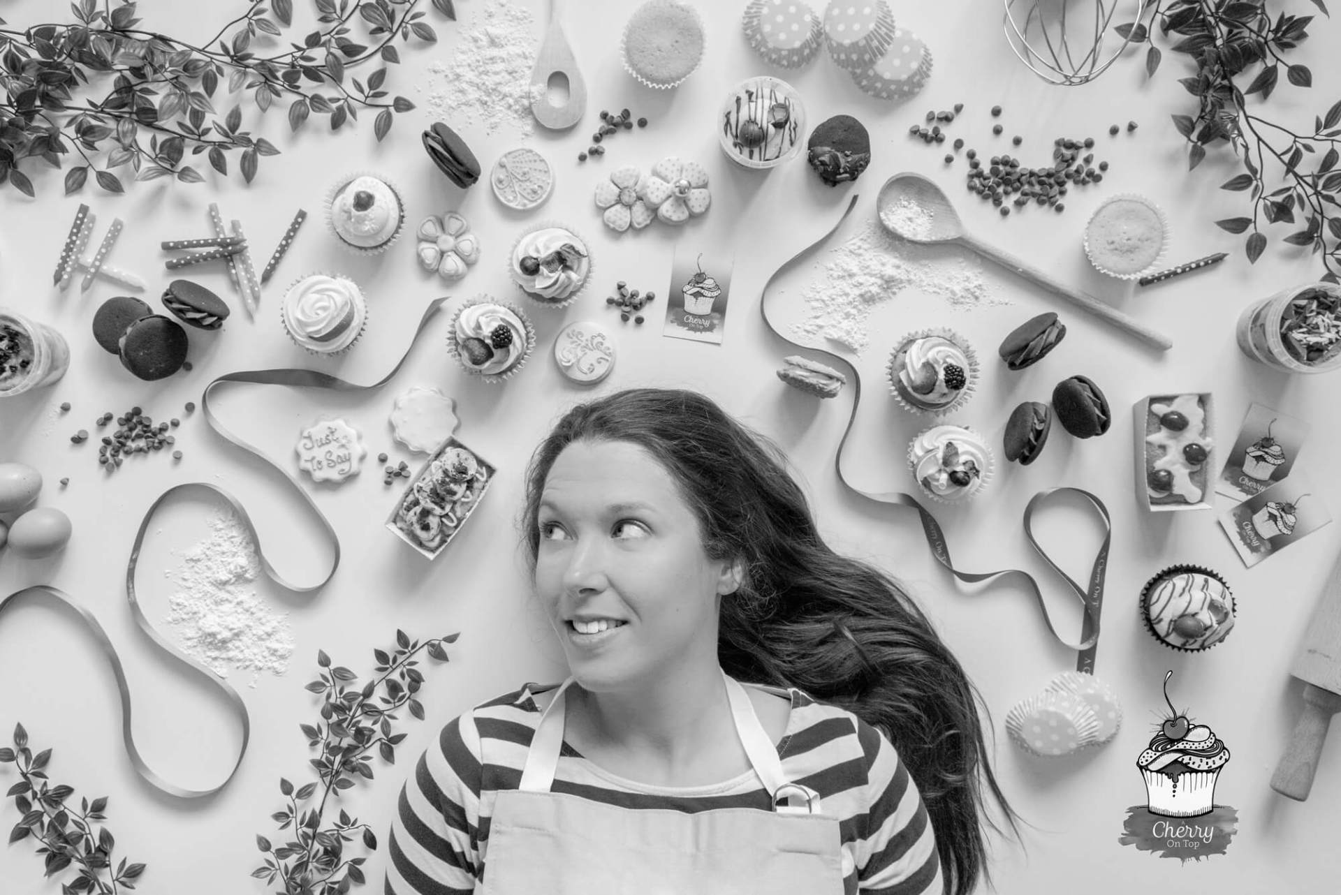 Woman surrounded by pastries and baking utensils looking up, in a kitchen.