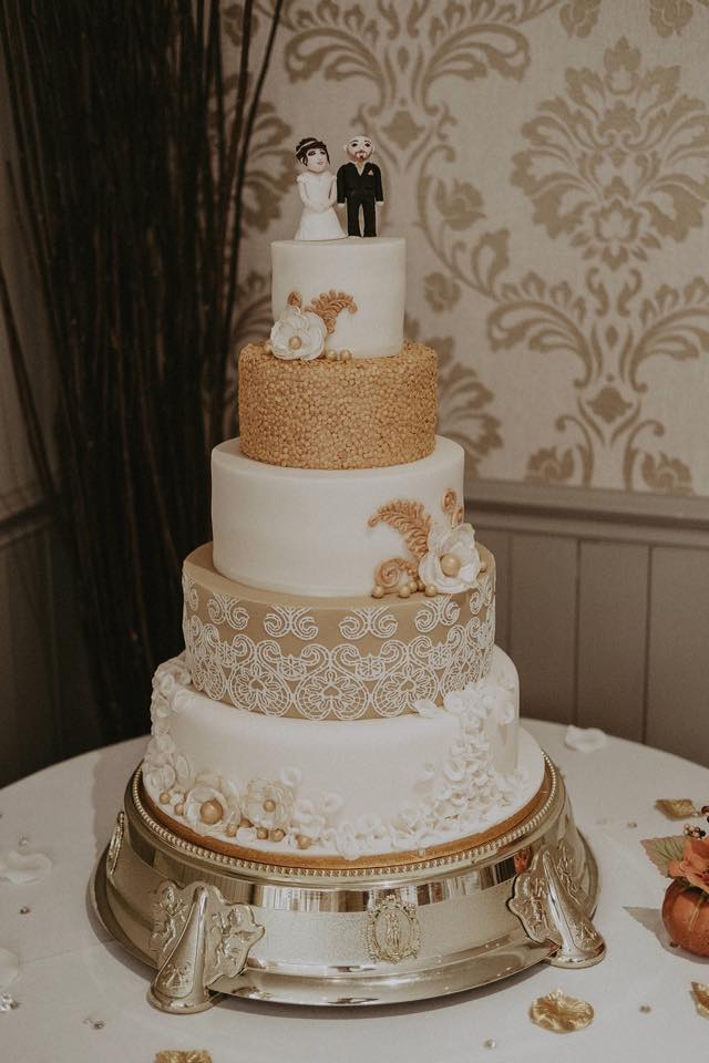 Five-tiered wedding cake with gold and white decorations, topped with bride and groom figurines on a silver cake stand.
