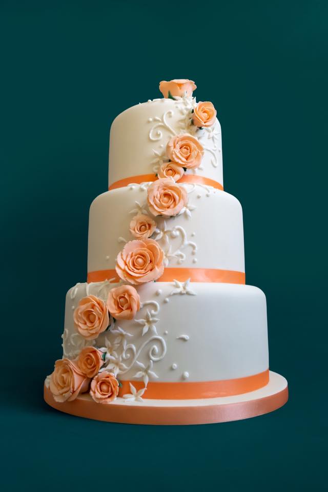 Three-tiered white cake with orange roses and ribbon on a teal background.