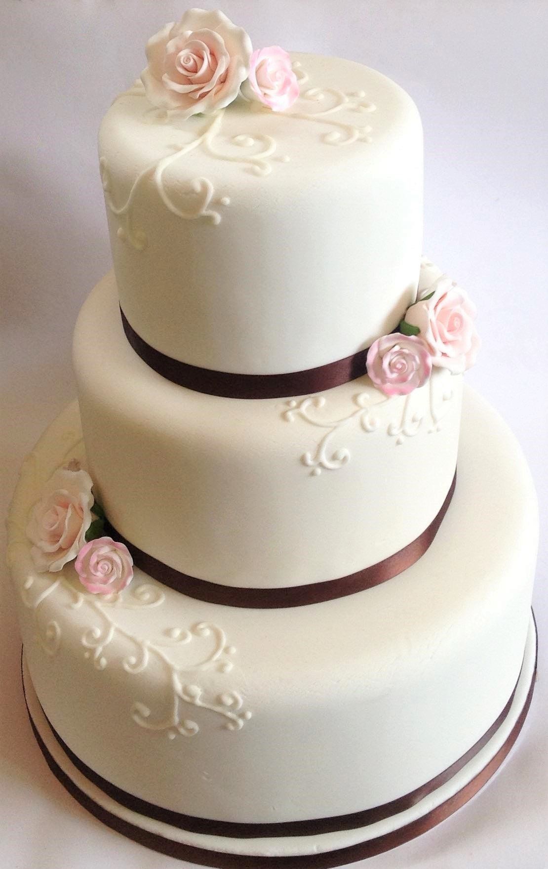 Three-tiered white wedding cake with pink roses and brown ribbon accents.