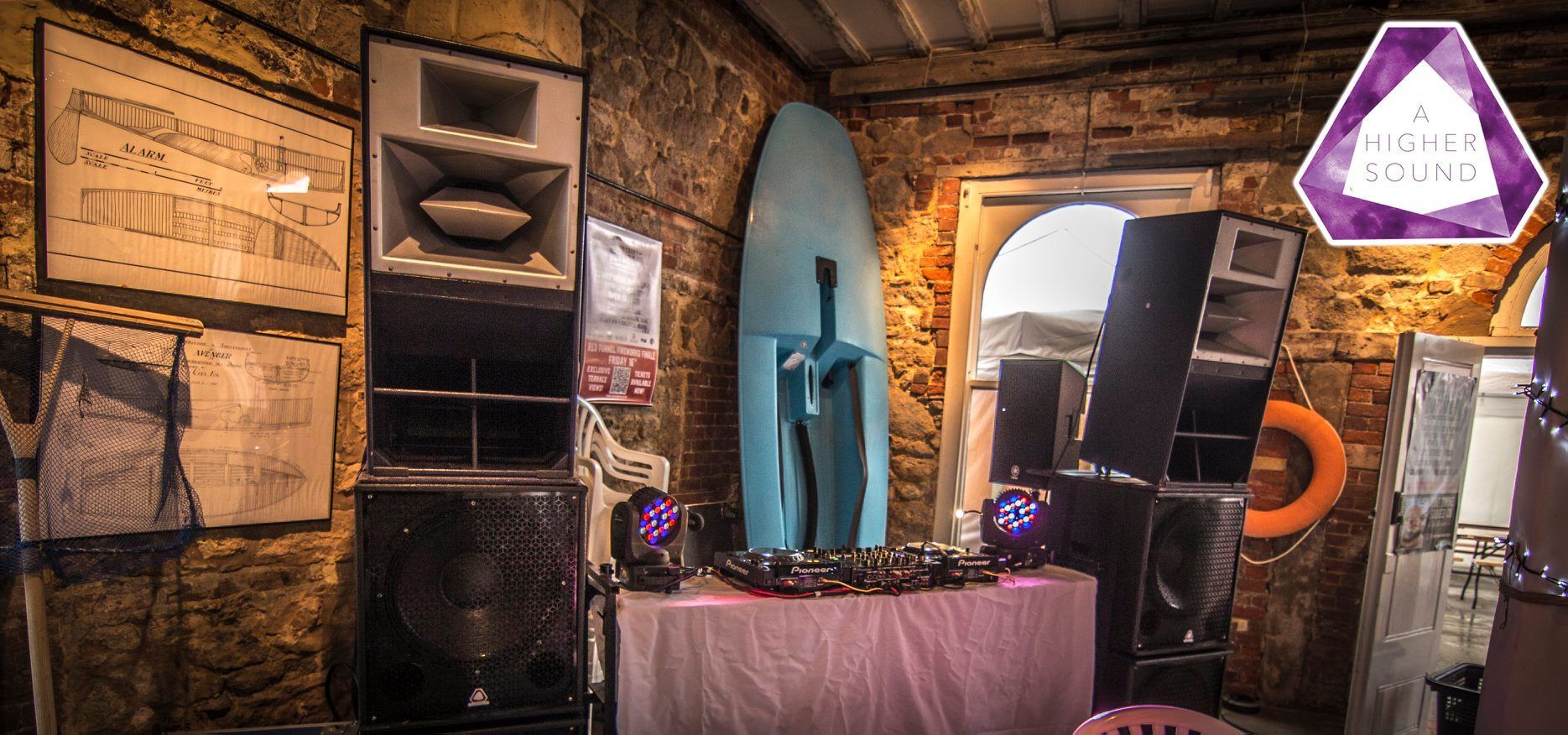 A DJ setup in a rustic room. Blue surfboard backdrop. Speakers, lights, a table.