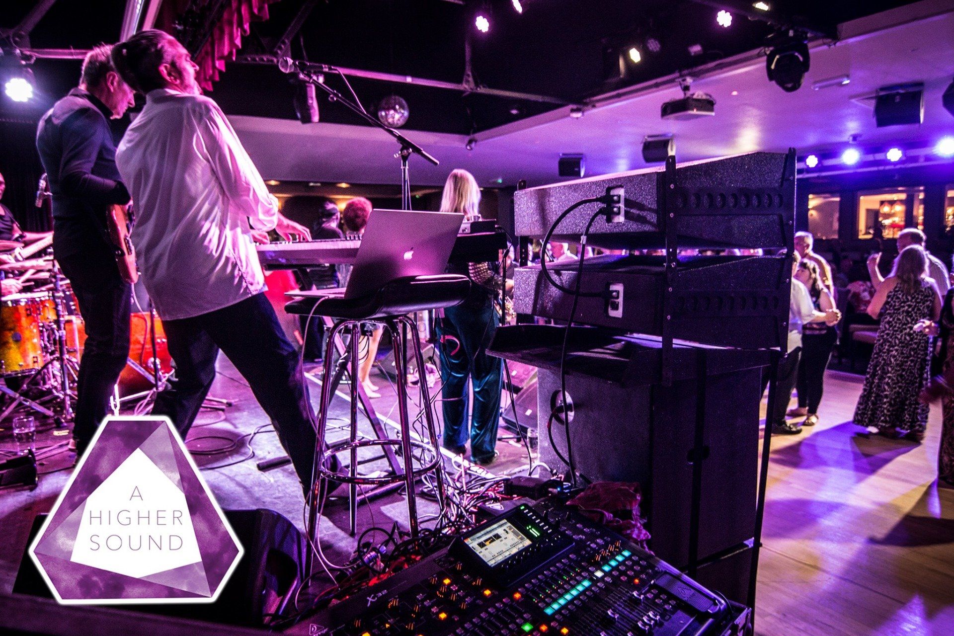 Band performing on stage with mixing console in foreground, audience dancing. Stage lit with purple and white.