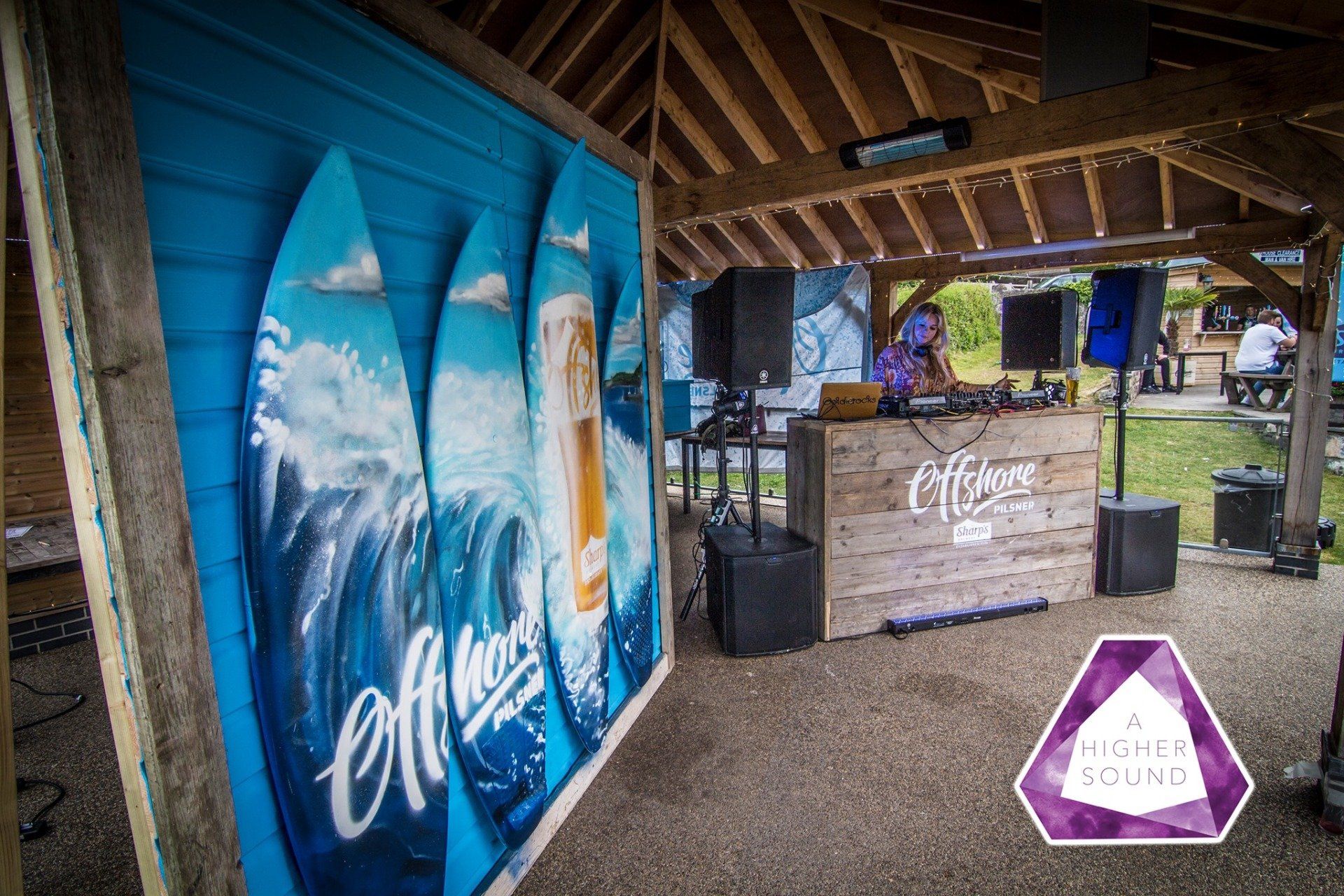 Outdoor DJ booth with wood paneling, surfboards, and music equipment. Purple and blue accents.
