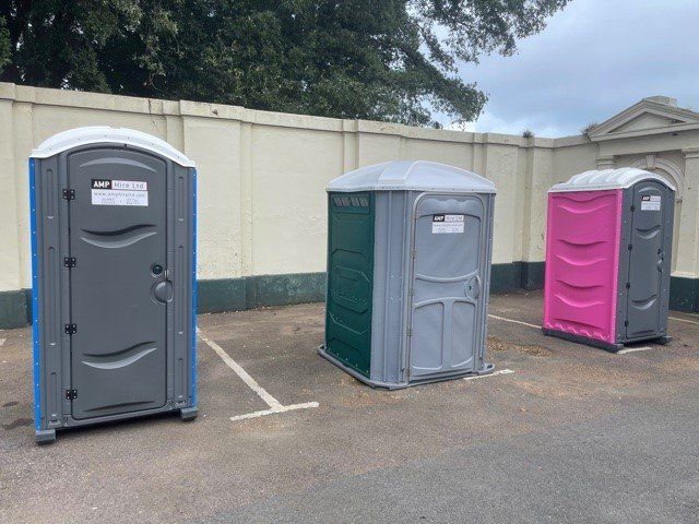 Three portable toilets of different colors in a parking lot against a light-colored wall.