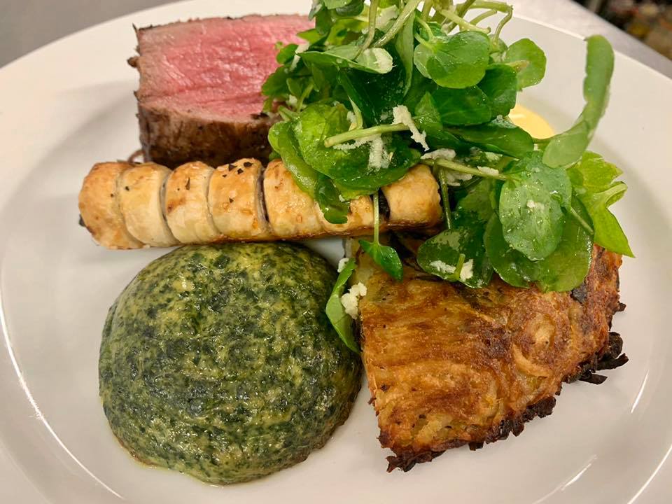 Plate of food with a medium-rare steak, puff pastry, potato cake, spinach, and watercress.