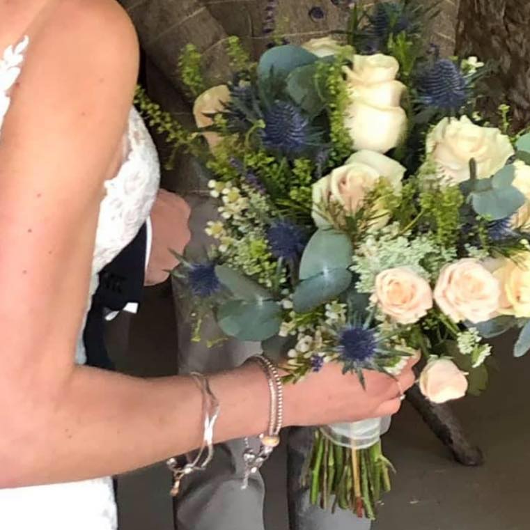 Bride holding a bouquet of white roses and blue thistles; man in suit standing beside her.
