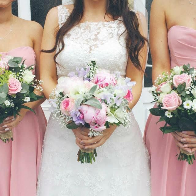 Bride in white lace dress, flanked by bridesmaids in pink dresses, holding flower bouquets.