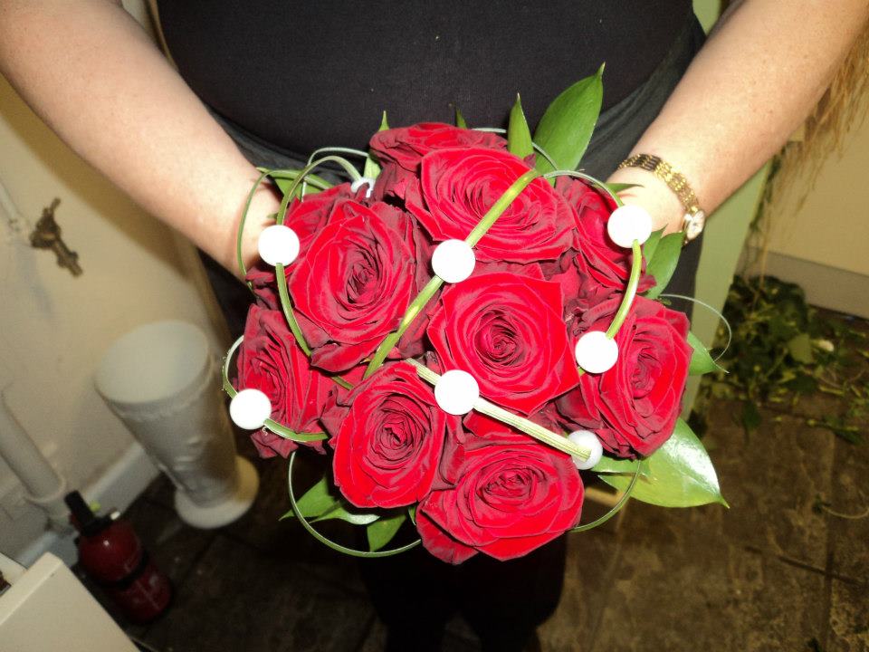 Person holding a bouquet of red roses, decorated with white beads and green stems.