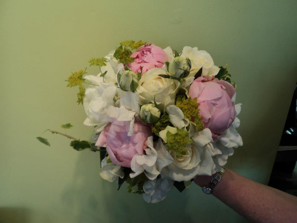 Hand holding a round bouquet of pink and white peonies and greenery against a green wall.