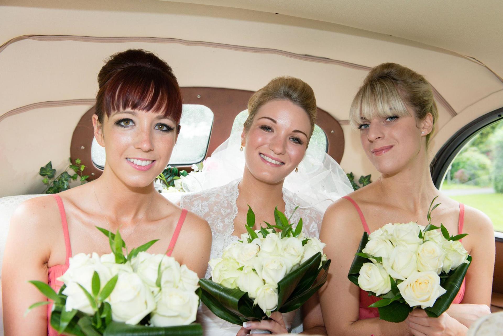 Bride and bridesmaids in a car, holding white rose bouquets. All smiling, wearing wedding attire.