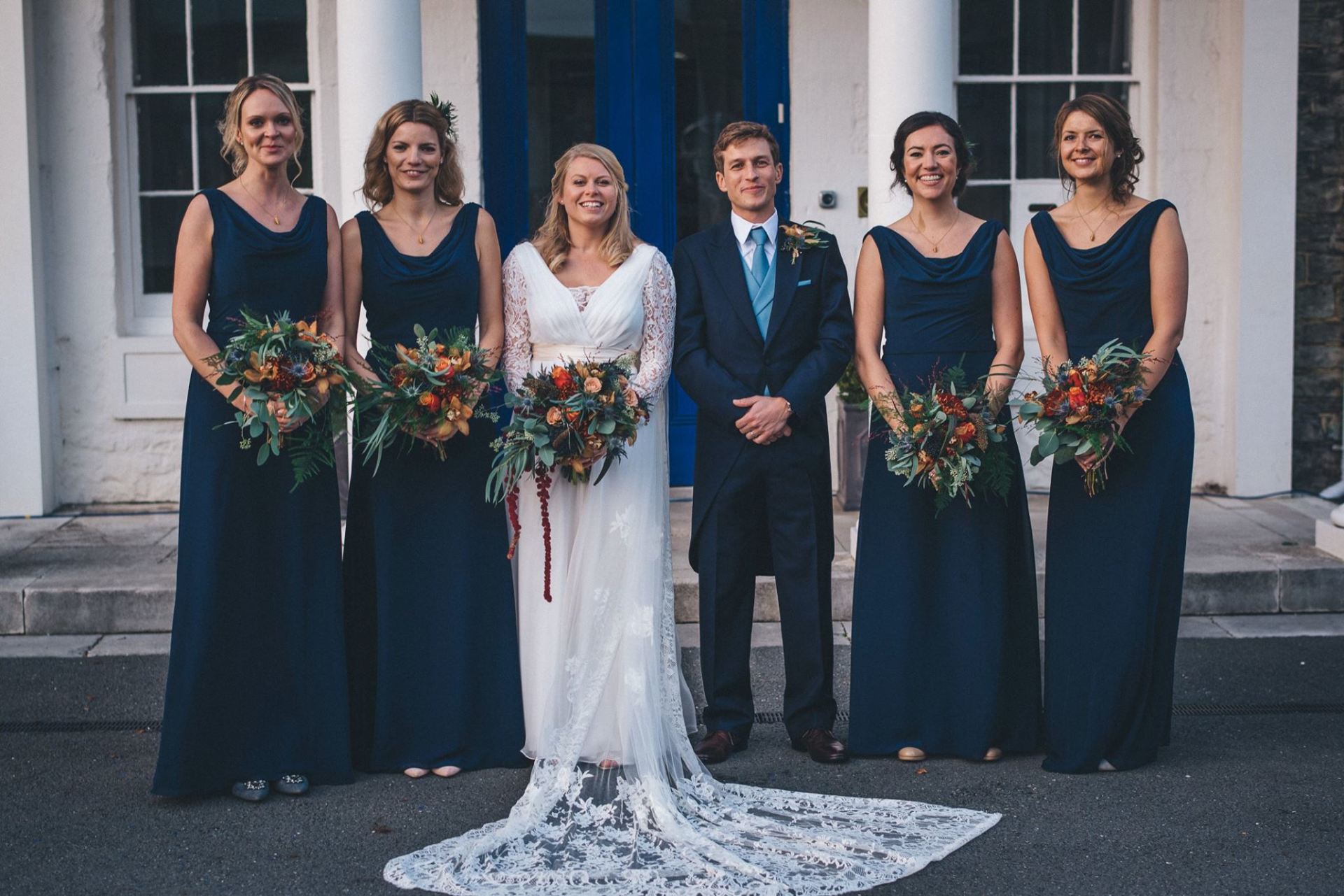 Wedding party posing outside; bride in lace dress, bridesmaids in navy dresses, groom in suit, holding bouquets.