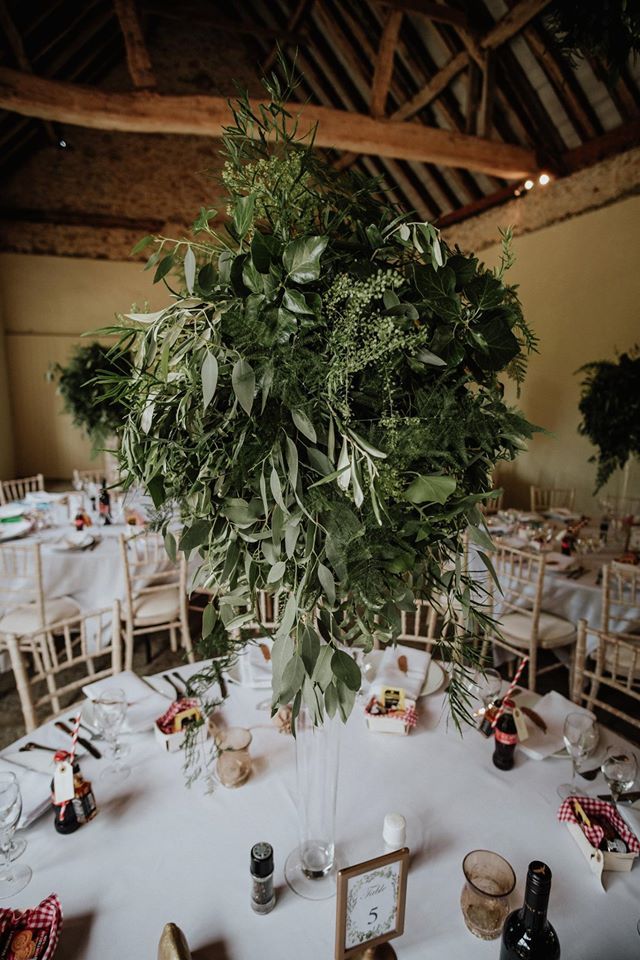 Wedding reception table with large green floral centerpiece and place settings, in a rustic venue.