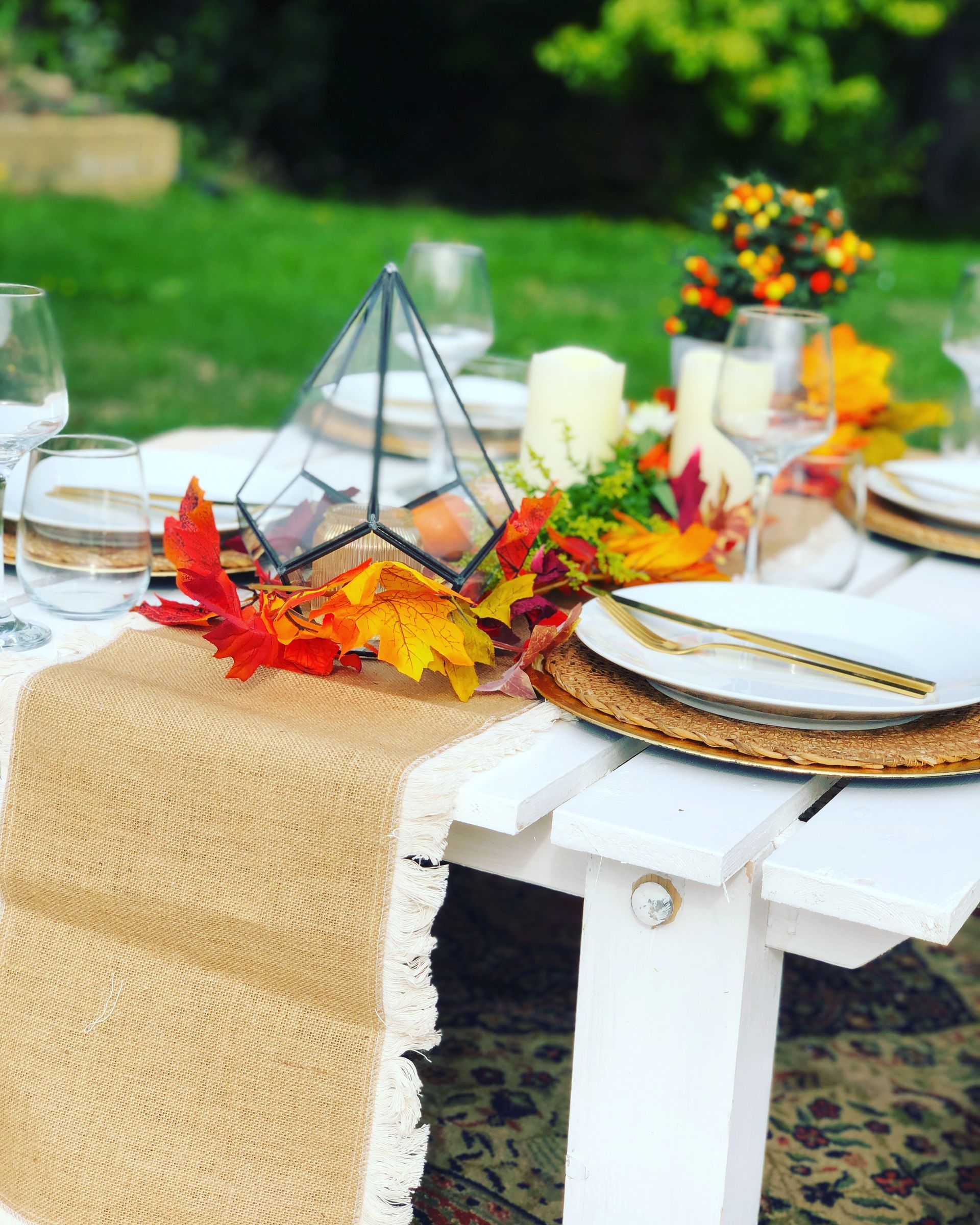 Outdoor autumn tablescape with burlap runner, fall foliage, and white dishes.