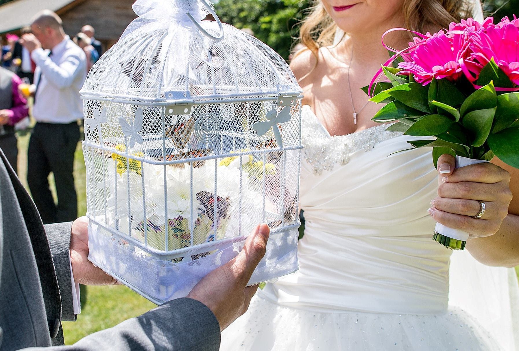 Bride in white dress holding pink bouquet and a birdcage with cupcakes at an outdoor wedding.