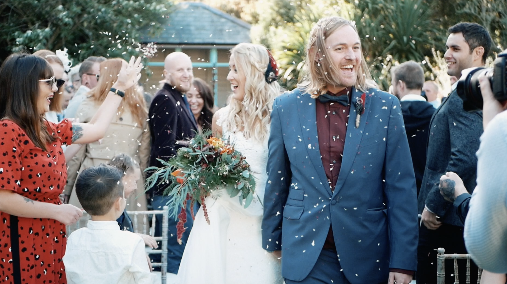 Newlyweds exit ceremony, confetti falling. Bride smiles, holding bouquet, groom laughs, wearing blue suit. Guests cheer.