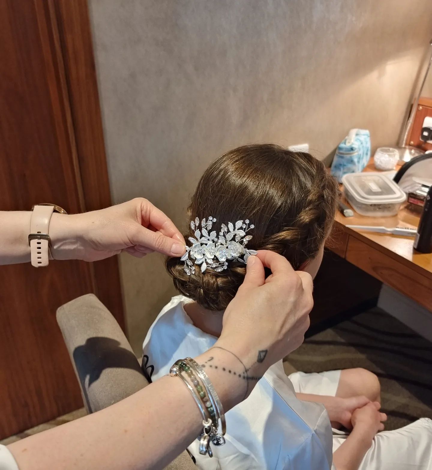 Hands securing a jeweled hairpiece in a woman's updo in a room with a table.