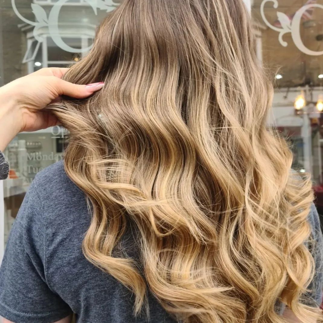 Woman with long, wavy brown and blonde highlighted hair; hand lifting some strands.