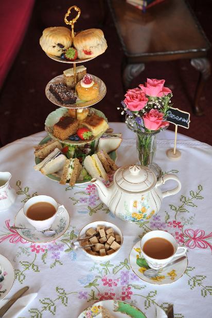 High tea table set with tiered food, flowers, and teacups, on a floral tablecloth.