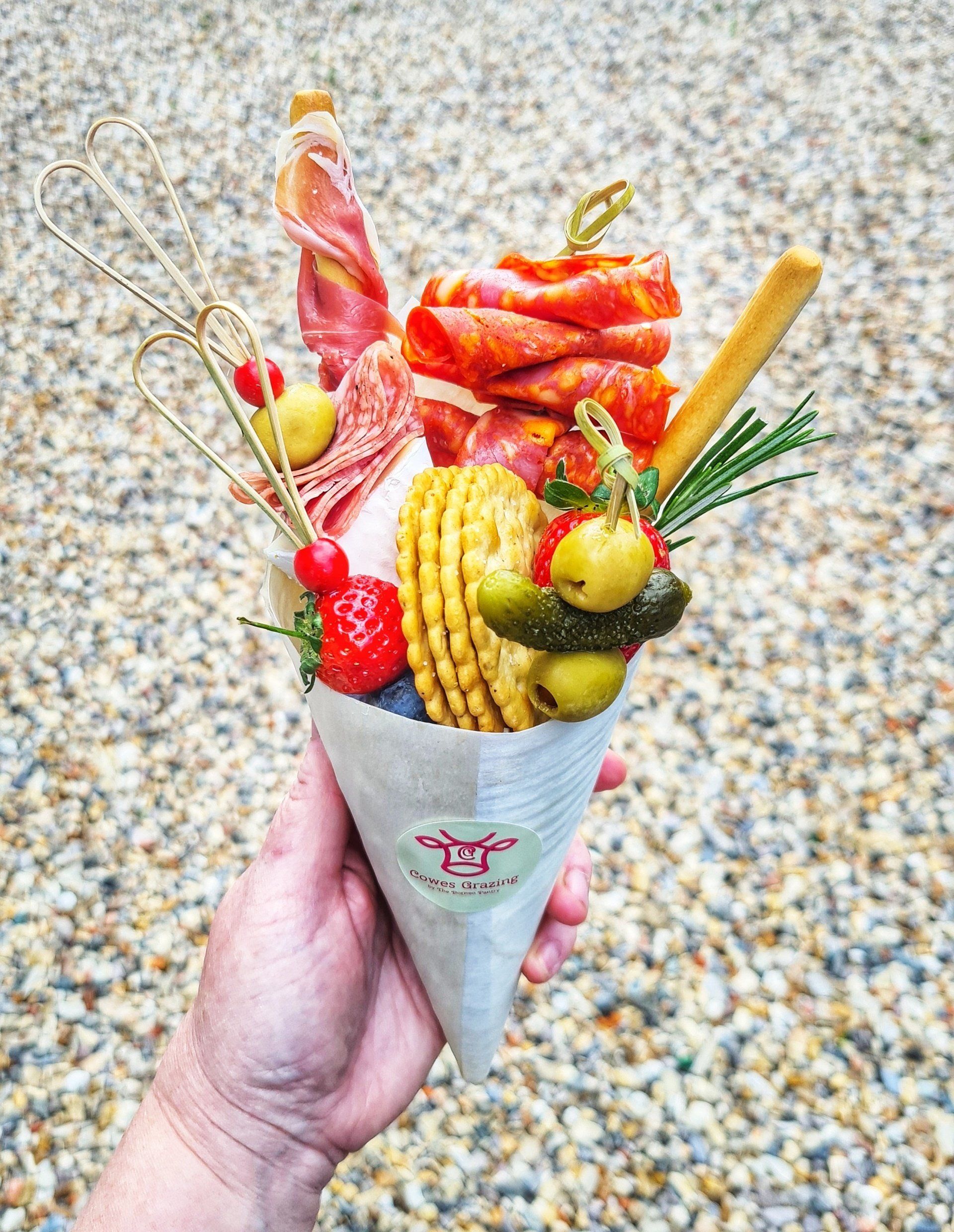 Hand holding a charcuterie cone with meats, cheeses, crackers, olives, and garnishes.