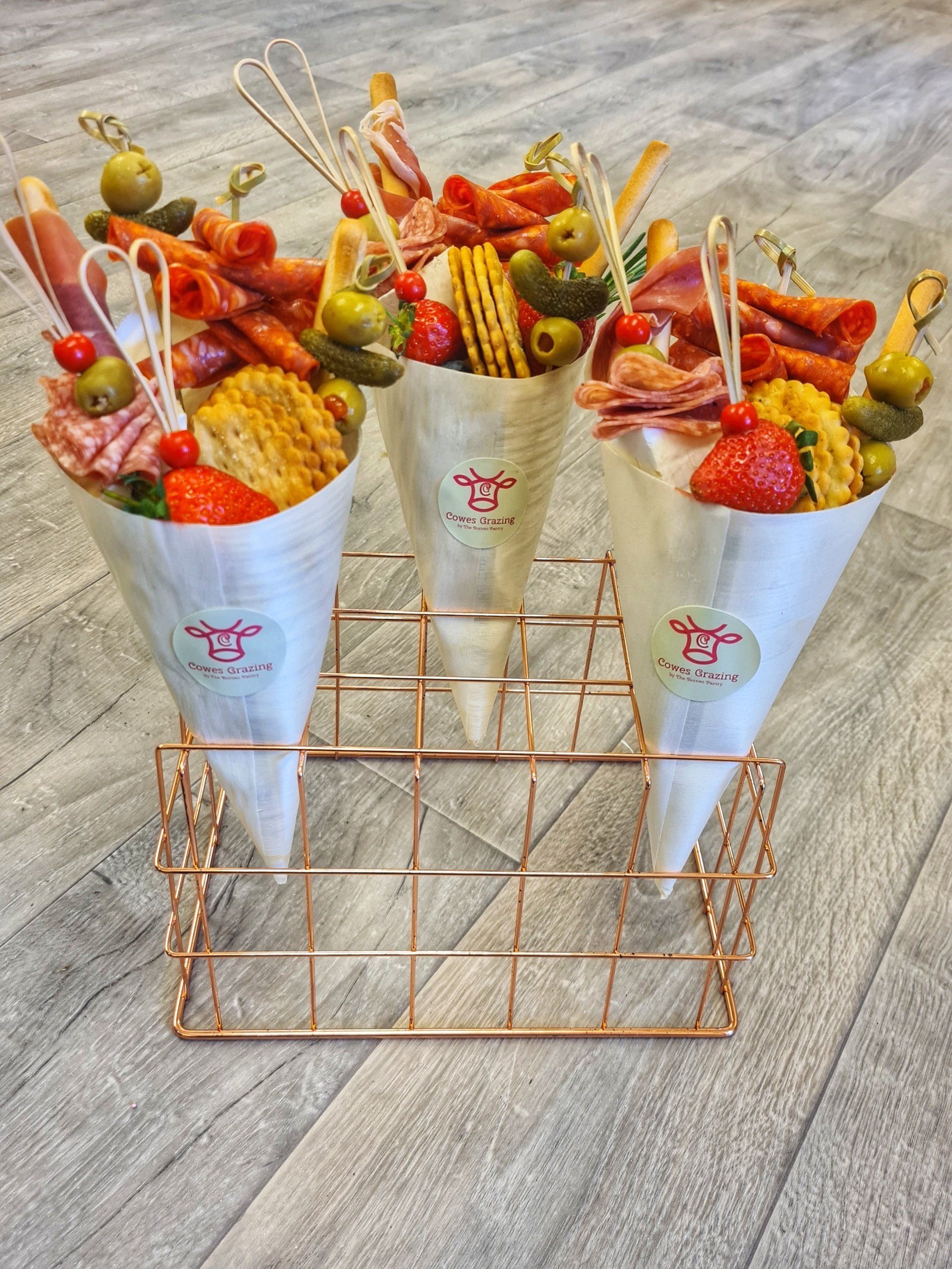 Three charcuterie cones with skewers, in a copper wire stand, on a wood-like surface.