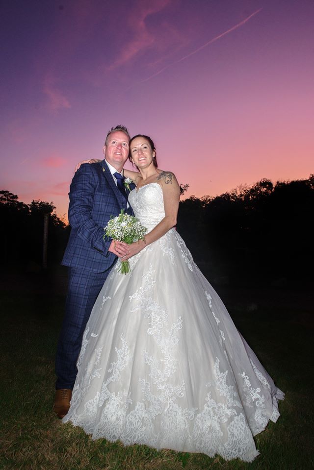 Wedding couple embraces outdoors at sunset. Groom in blue suit, bride in white gown. Pink and purple sky.