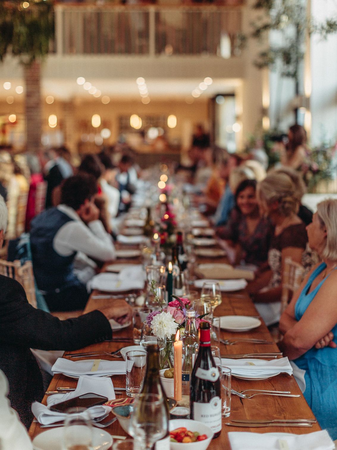 Long wooden dining table set for a wedding, with guests seated and smiling, candles and flowers.