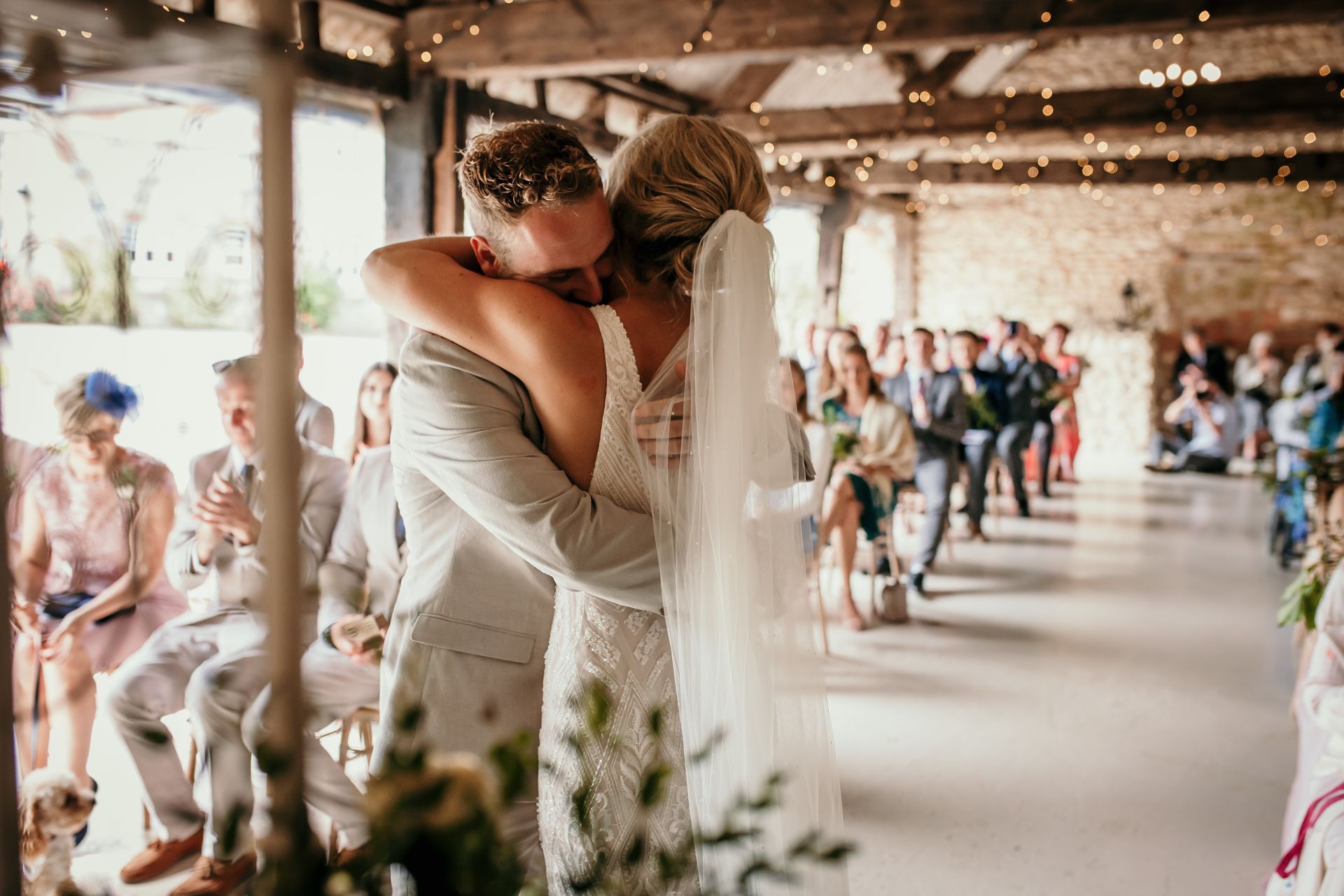 Newlyweds embrace, kissing at their wedding ceremony inside a barn, guests seated in background.