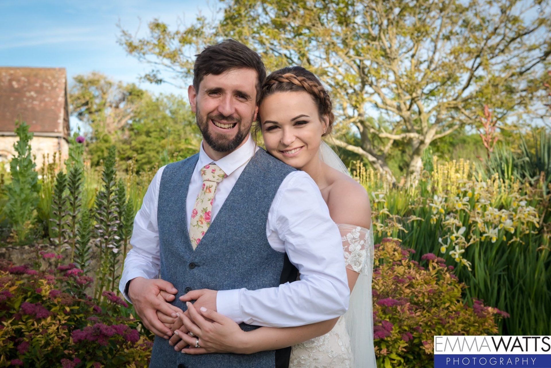 Wedding couple embracing outdoors, man in vest, woman in white dress, sunny day.