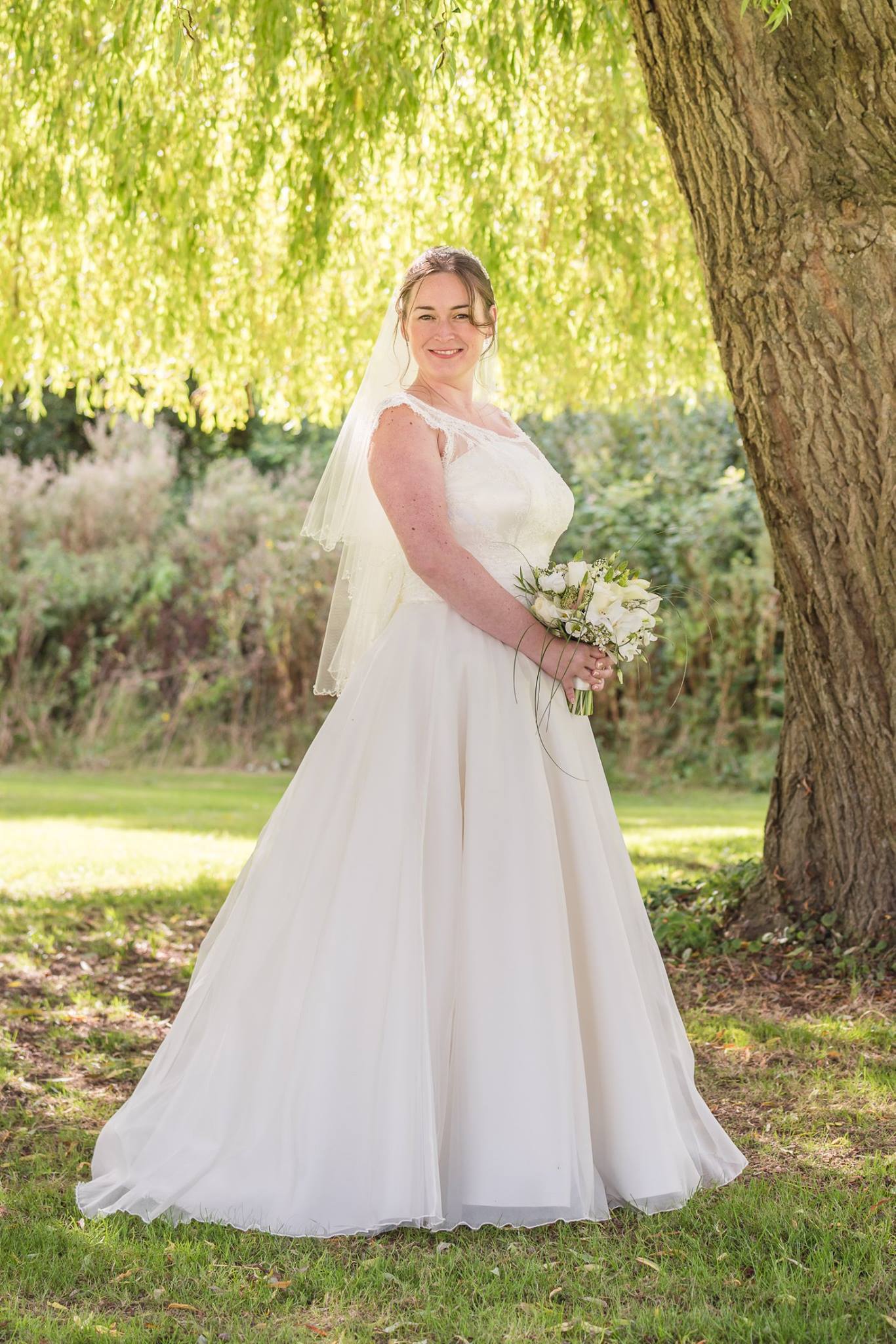 Bride in white dress, veil, and bouquet, standing under a willow tree in a sunny outdoor setting.