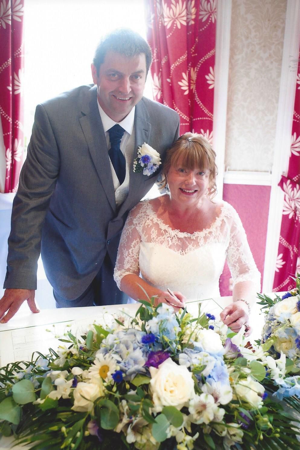 Couple signing a marriage certificate. Man in grey suit, woman in white dress, floral arrangement.