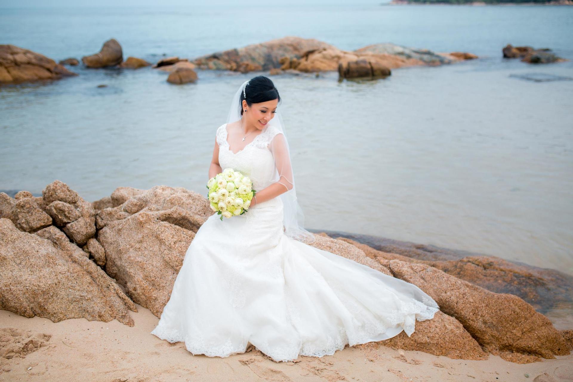 Bride in white dress holding bouquet sits on rocks by the ocean.