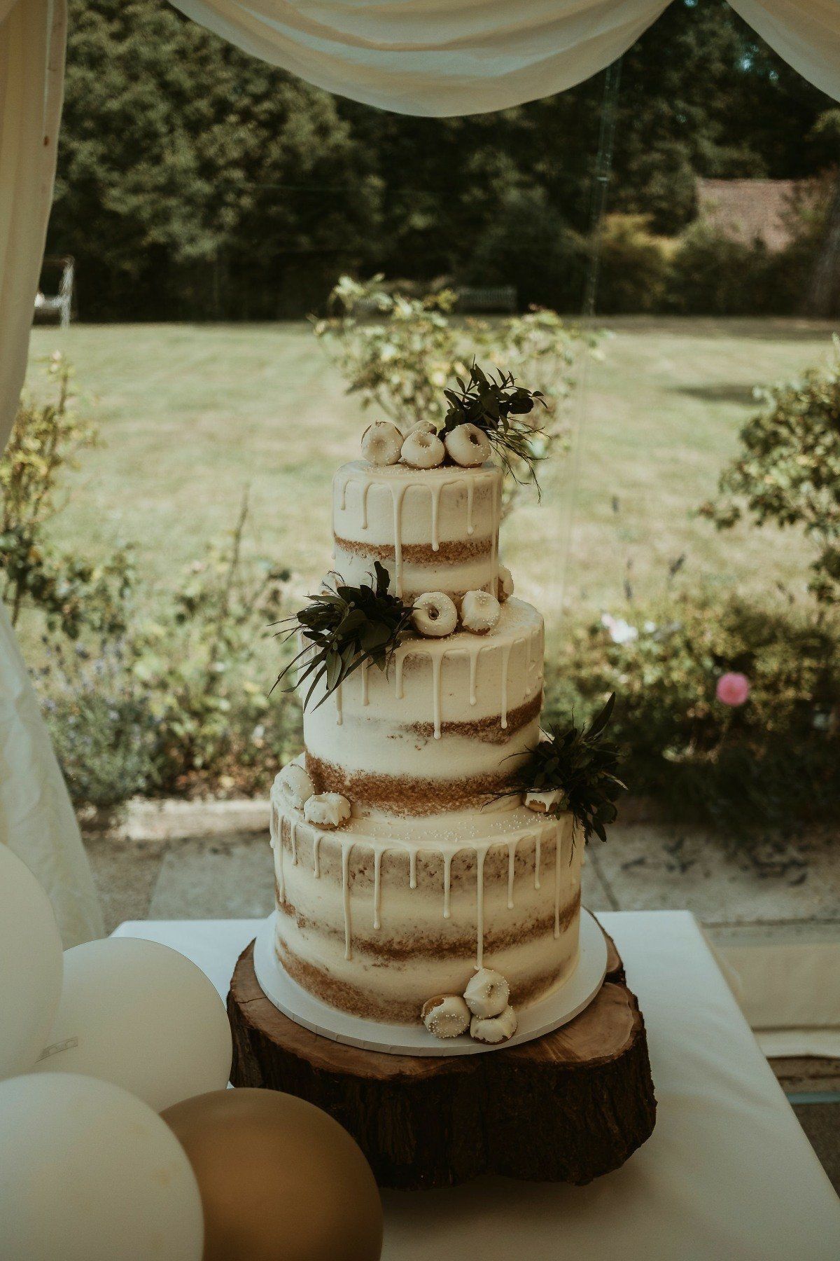 Four-tiered cake with dripped frosting, donuts, and greenery, on a wood slice. Outdoors, near a window.