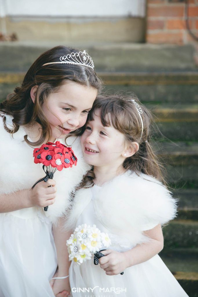 Two young girls in white dresses and fur wraps, holding bouquets, looking at each other, smiling.