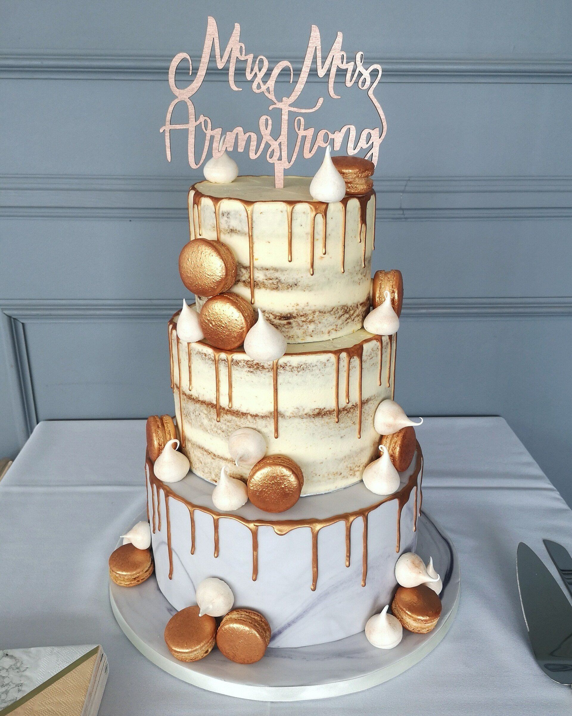 Three-tiered wedding cake with gold drip, macarons, and meringue. Topper reads