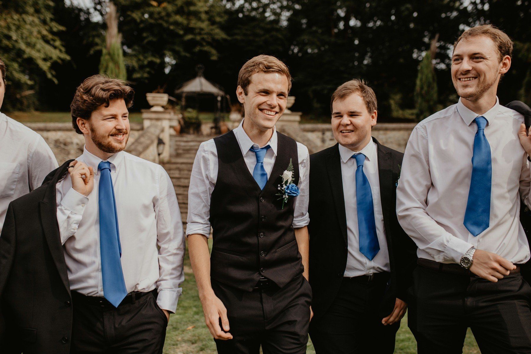 Groomsmen in suits and blue ties walking outdoors, smiling.