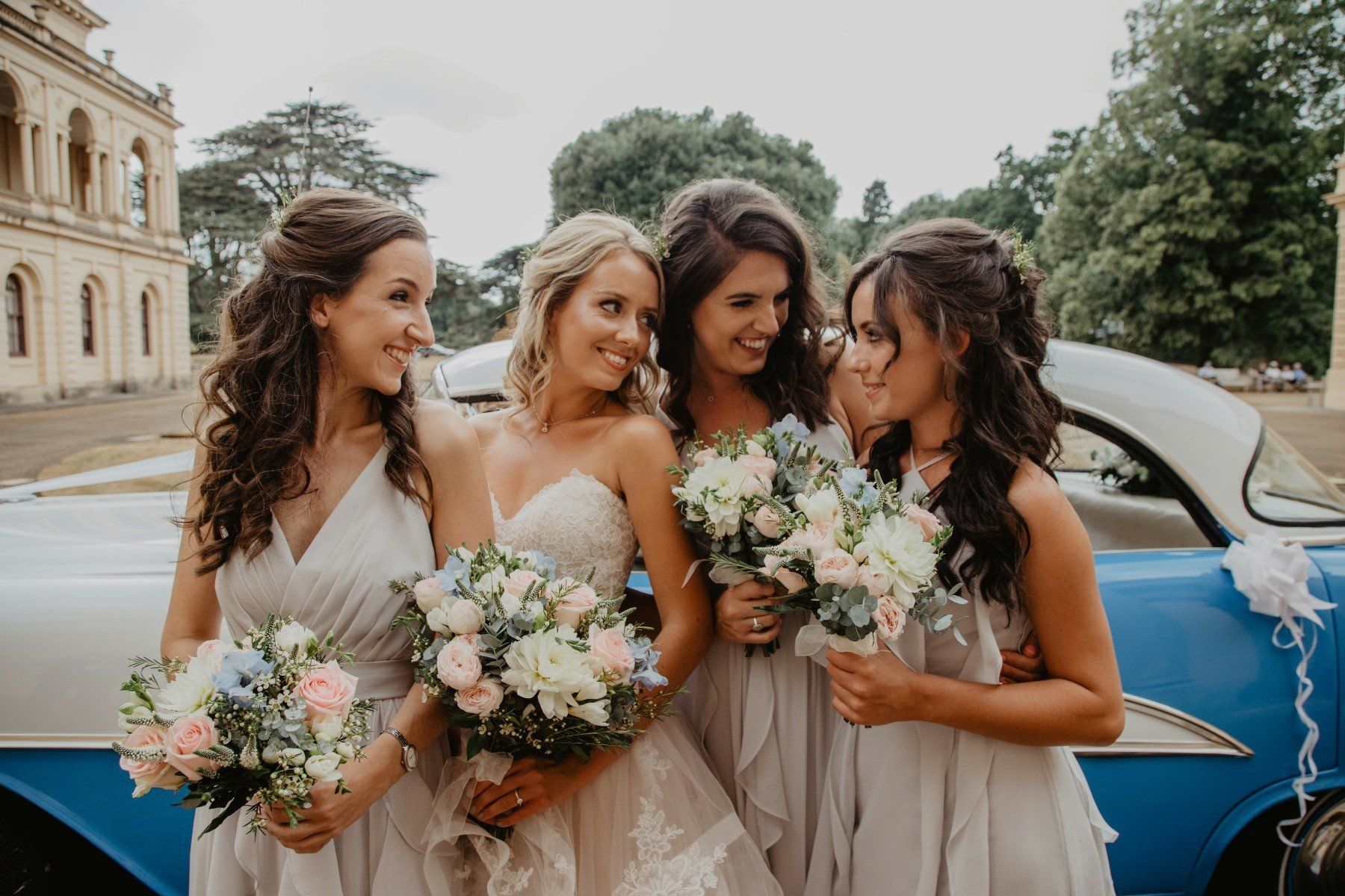 Bridesmaids and bride pose next to a vintage blue car, holding bouquets. They are smiling in a garden setting.