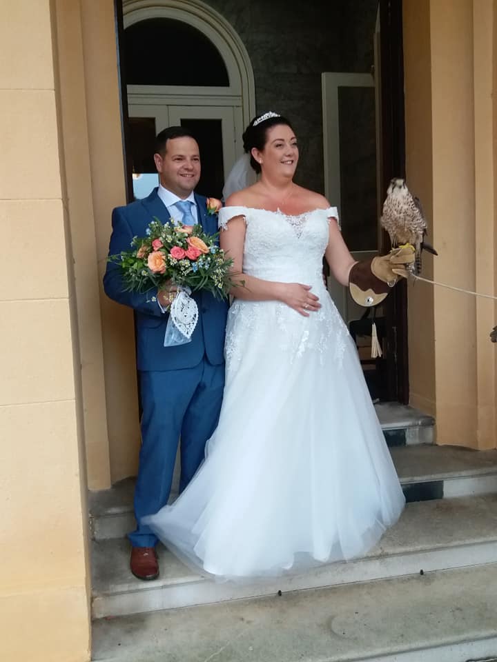 Newlyweds pose with a falcon. Man in blue suit holds bouquet, woman in white gown holds falcon, both smiling. Stone doorway.