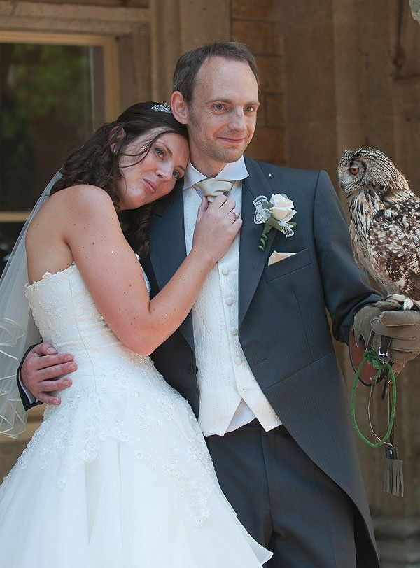 Bride and groom pose with owl at their wedding. The bride leans on groom, who holds the owl on his gloved hand.