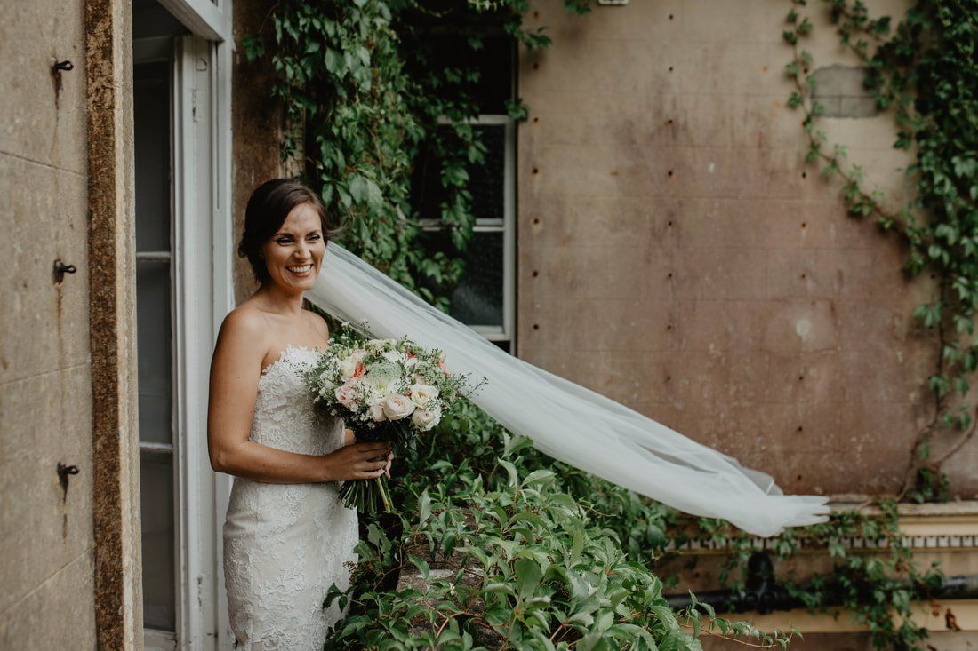 Bride in strapless lace dress smiles, holding bouquet, veil blowing near ivy-covered building.