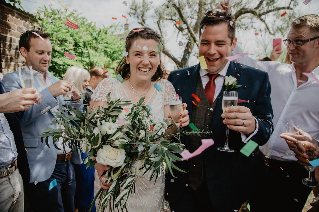 Newlyweds showered with confetti, holding champagne, smiling, outdoors.