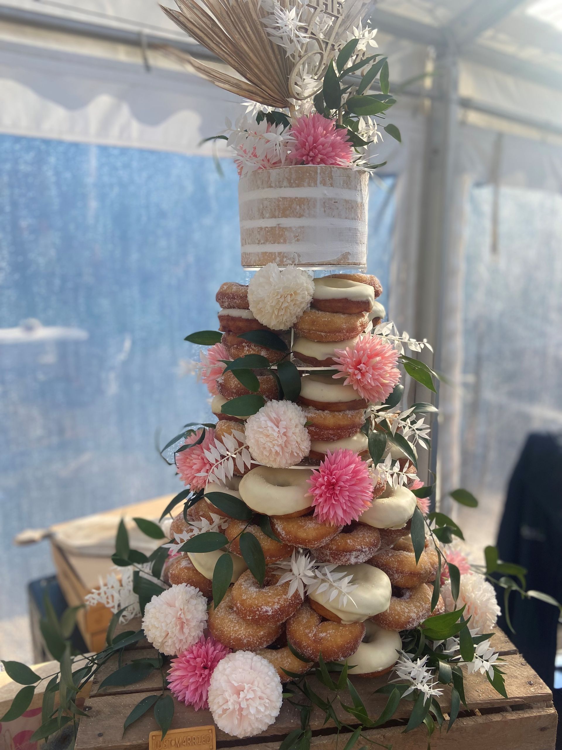 Donut cake decorated with pink flowers and greenery, topped with a small cake and wheat stalks.