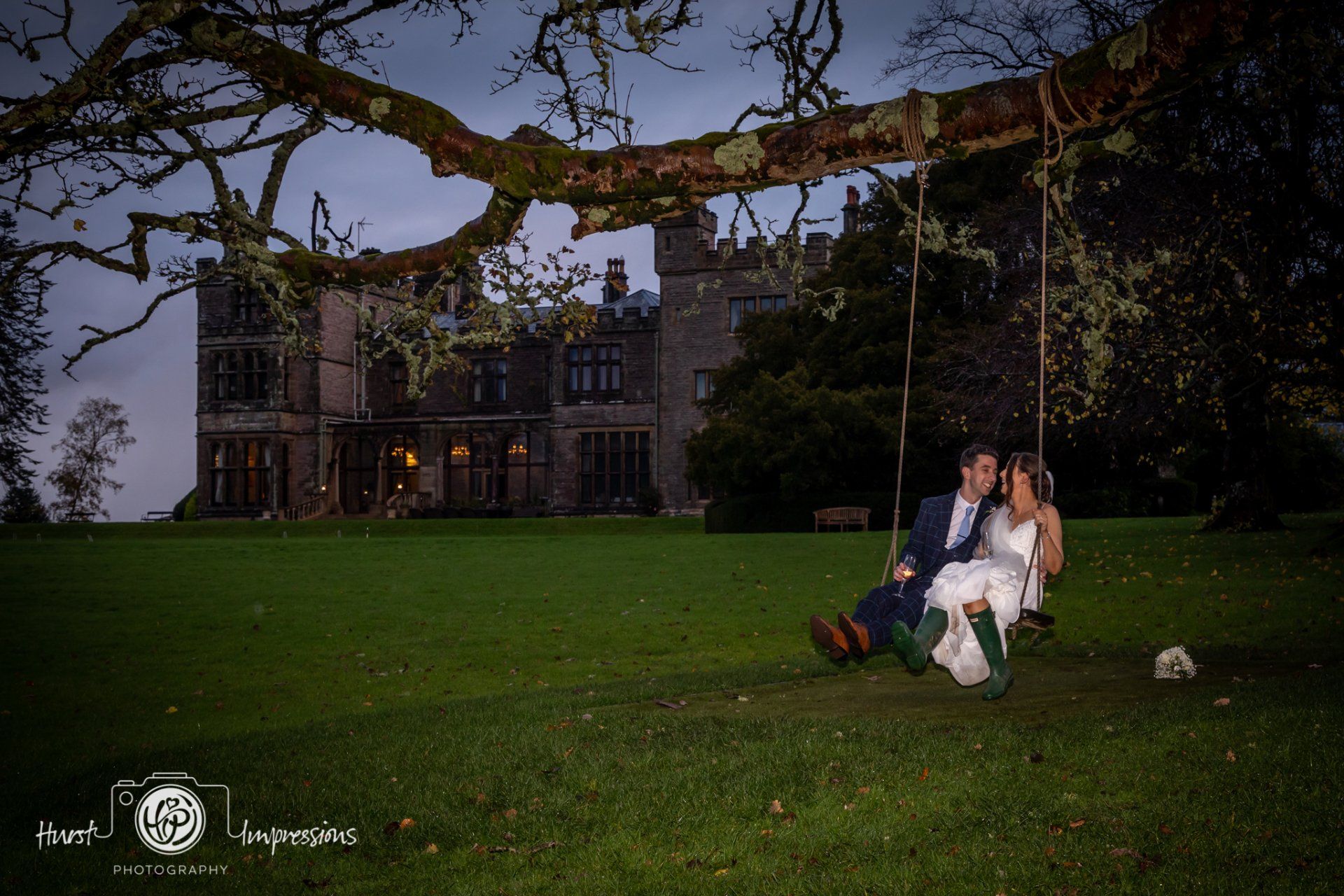 Couple on swing, castle in background. They are smiling, wearing wedding attire, and sitting on a swing under a large tree.